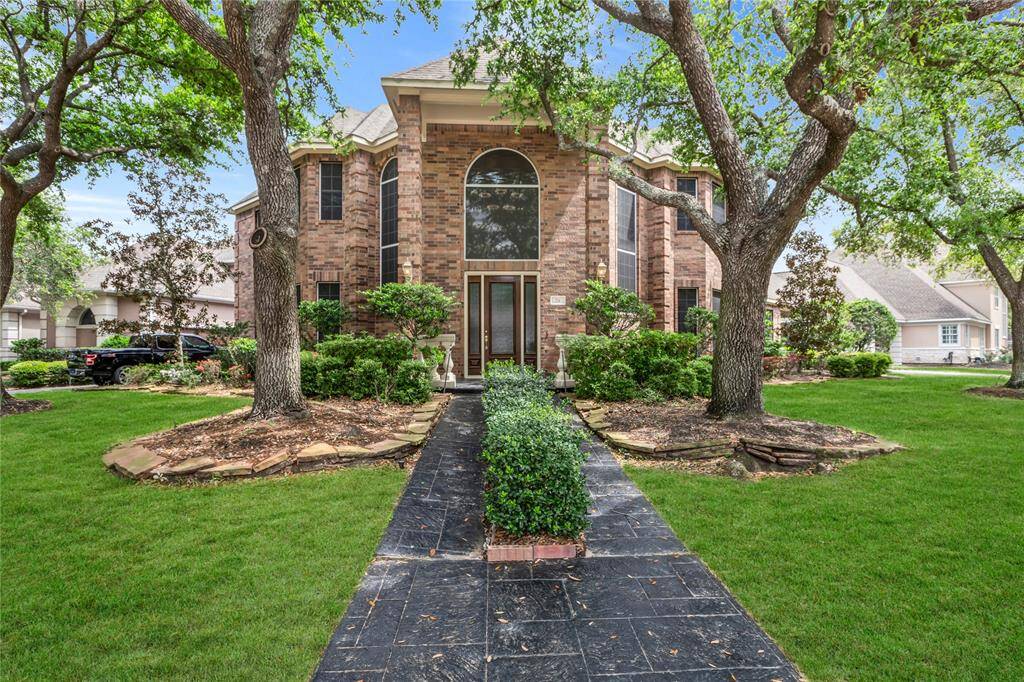 This elegant brick home features a grand entrance with large arched windows, surrounded by mature trees and well-maintained landscaping. The pathway leads through lush greenery, offering a welcoming and picturesque frontage.
