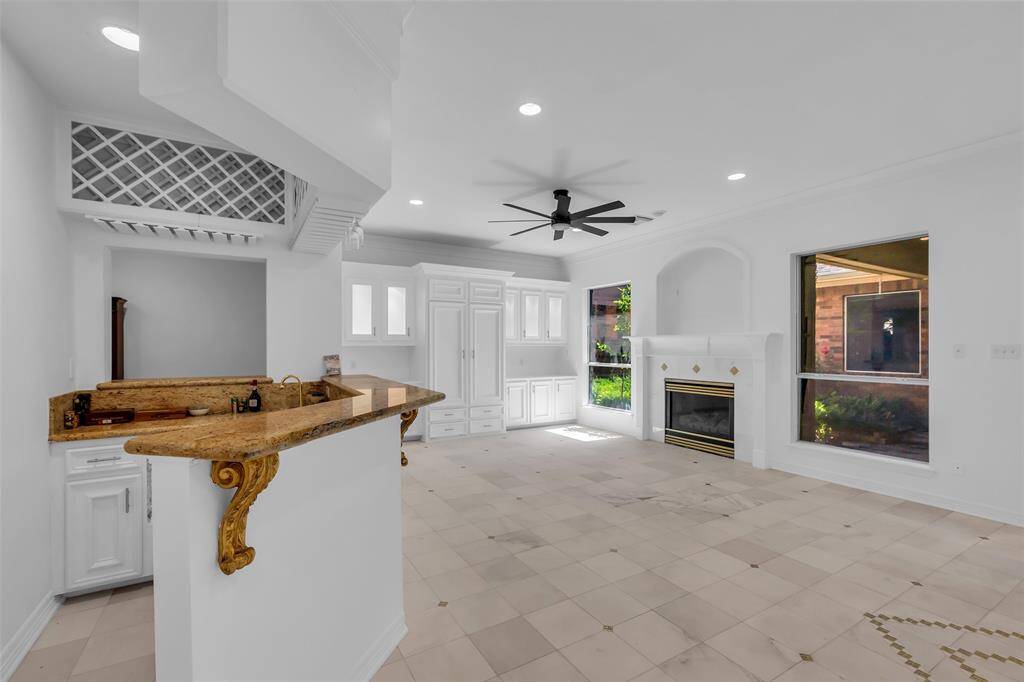 This spacious, bright living area features elegant tile flooring, a cozy fireplace, large windows for natural light, and a stylish ceiling fan. The open layout connects to a kitchen bar with decorative wood accents, perfect for entertaining.