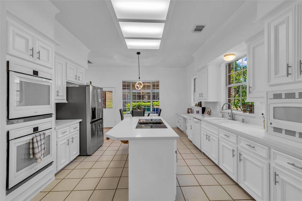 Kitchen with white cabinetry and ample counter space. Features include double ovens, a central island with a stovetop, and large windows offering natural light. Adjacent dining area enhances the open layout.