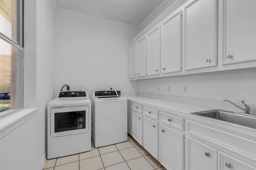 This photo shows a spacious laundry room with a washer and dryer, ample cabinetry for storage, a countertop for folding, and a sink. It features a window for natural light and has a clean, white finish.
