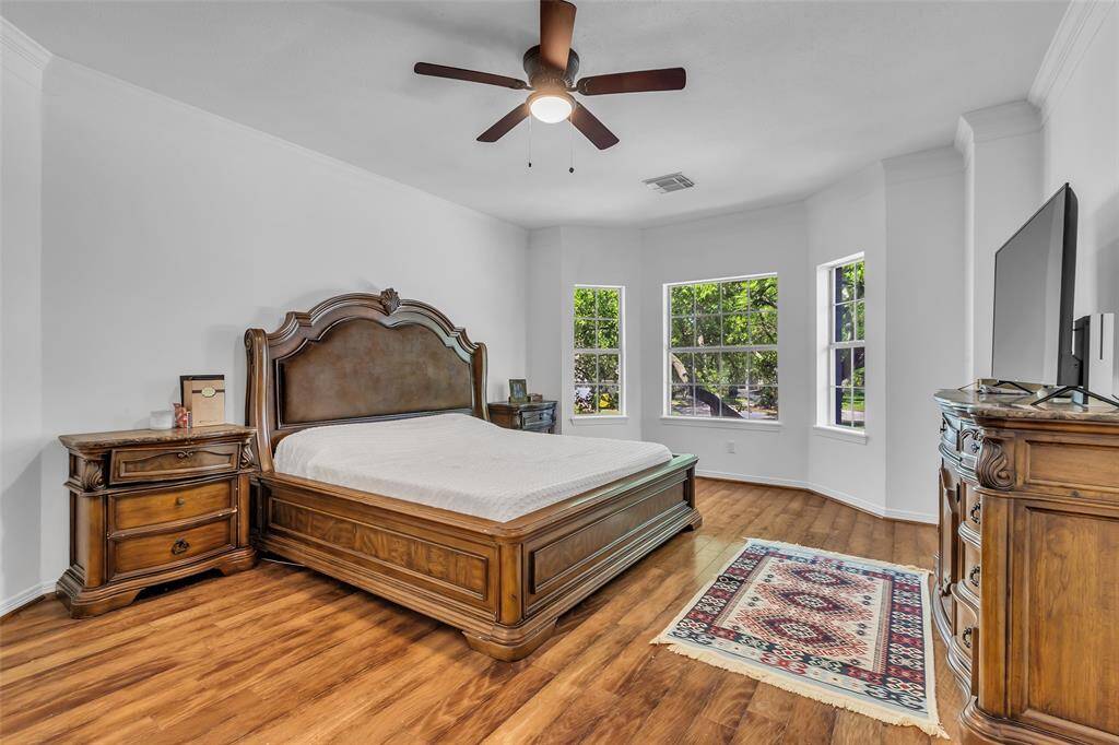 his spacious bedroom can accommodate a large bed & multiple pieces of furniture. The room has warm wood flooring, a ceiling fan, and a TV mount, creating a cozy and inviting atmosphere.