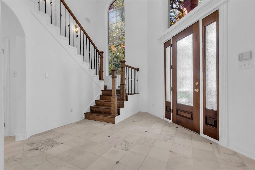 This two story entryway features a grand staircase with wooden steps and wrought iron railing. It has high ceilings, a large arched window for natural light, and elegant double doors with frosted glass panels, creating an inviting and spacious atmosphere.