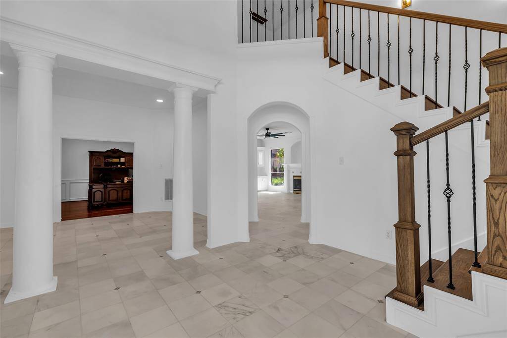 The photo showcases an elegant foyer with marble flooring, decorative columns, and a wooden staircase with wrought iron railings leading to the bedrooms upstairs.