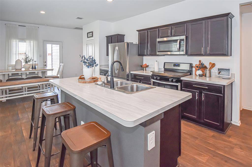 This modern kitchen with dark cabinetry and stainless steel appliances. It features a central island with a sink and bar stools. The space is bright with natural light, offering a welcoming and functional layout.