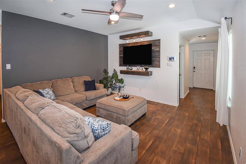 A wall-mounted TV with a wooden accent shelf adds modern flair. The room has recessed lighting and a ceiling fan for comfort.