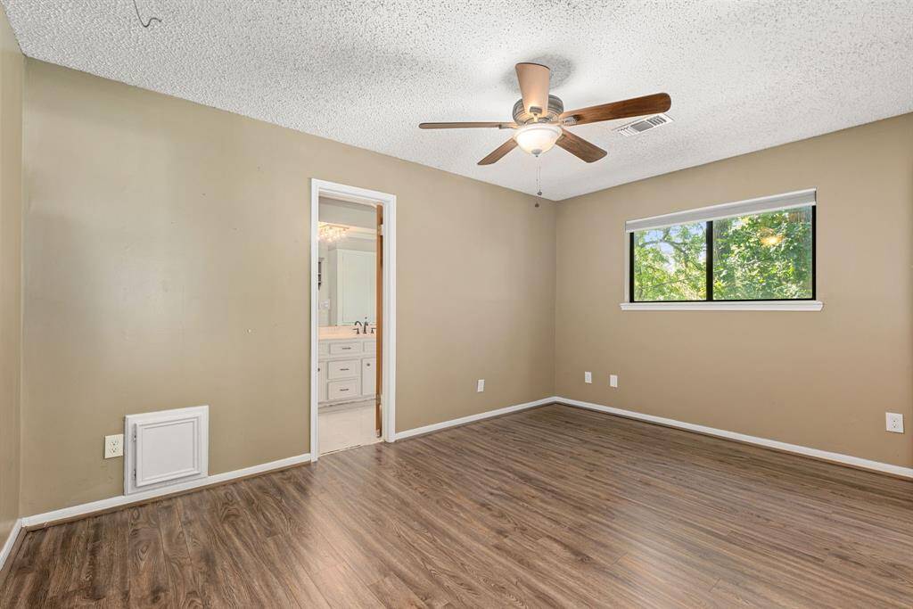 This bedroom features wood flooring, a ceiling fan, and a large window offering natural light. An adjoining en-suite bathroom in view.