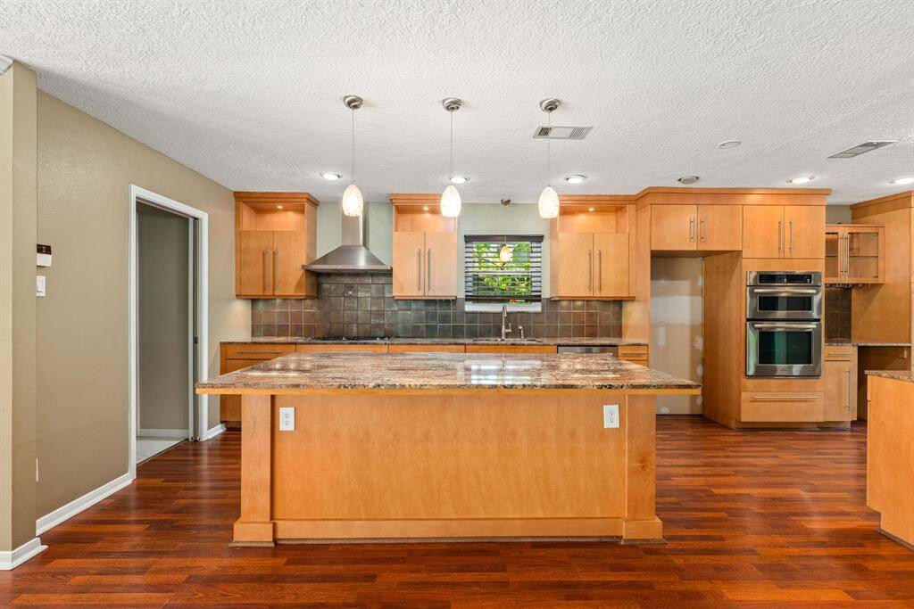 Large central island with granite countertops. Featuring under cabinet lights, recessed lighting, pendant fixtures, and rich hardwood flooring, creating an inviting space for cooking and entertaining.