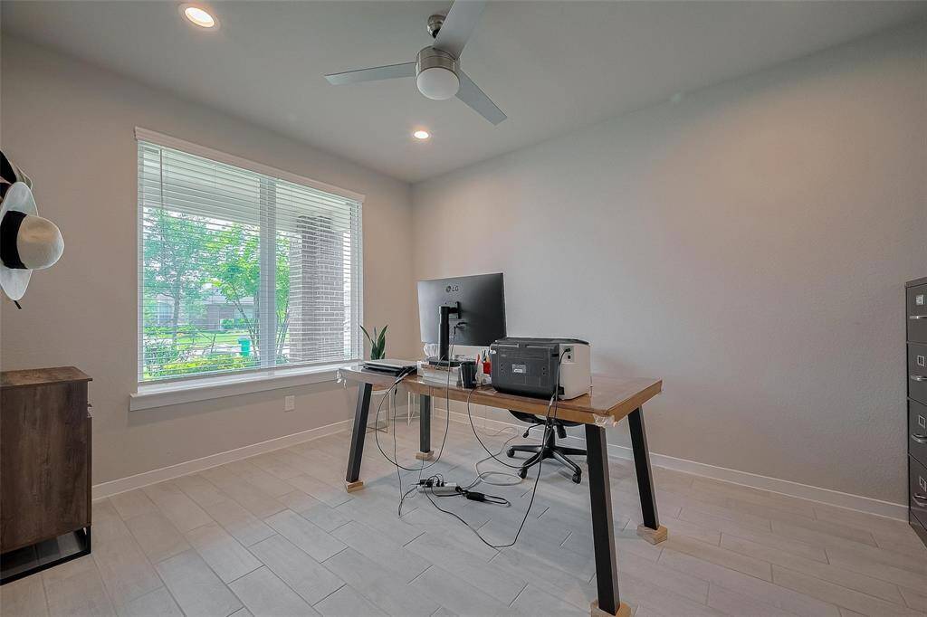 This photo shows a bright, modern home office with a large window providing natural light. The room features neutral walls, a ceiling fan, and tiled flooring. A simple desk with a computer and printer is set up, making it ideal for remote work or study.