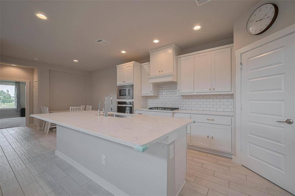 Modern kitchen with white cabinetry, subway tile backsplash, and a large island with a sink. Equipped with built-in appliances and recessed lighting. Open layout with light-colored flooring and a visible dining area.