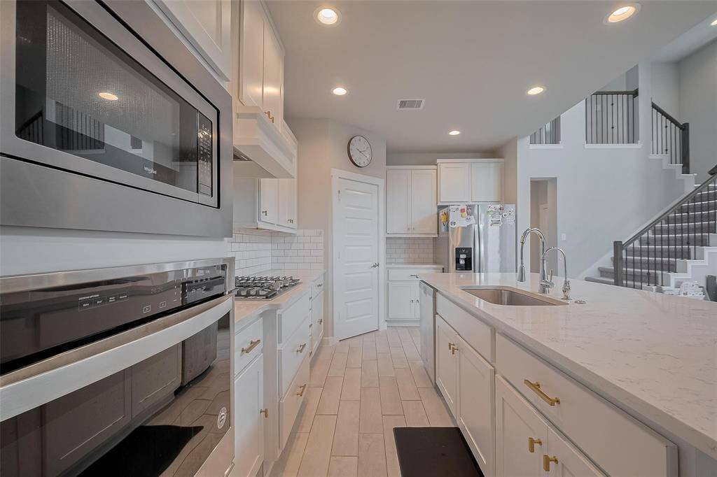 This modern kitchen features sleek stainless steel appliances, bright white cabinetry, and a spacious island with a sink. It's complemented by a stylish tile backsplash, ample lighting, and an open layout that seamlessly connects to the living area with a staircase.