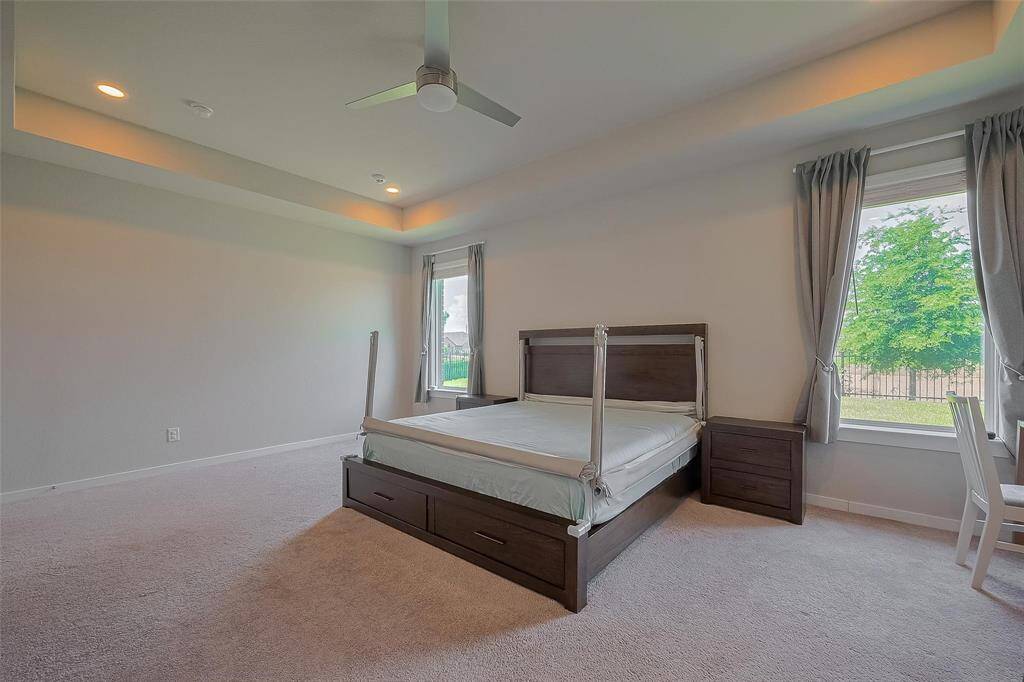 This spacious bedroom features a tray ceiling with recessed lighting, a ceiling fan, and large windows with curtains, providing plenty of natural light.