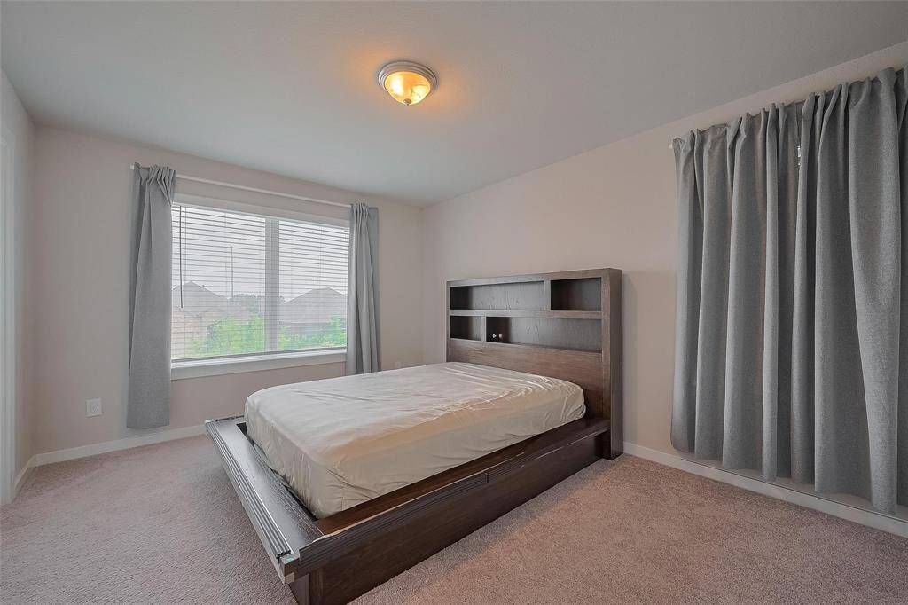 This photo showcases a bright, airy bedroom with a large window, neutral walls, and carpeted flooring. The room features a modern wooden bed frame with built-in shelving on the headboard, and simple gray curtains add a touch of elegance.
