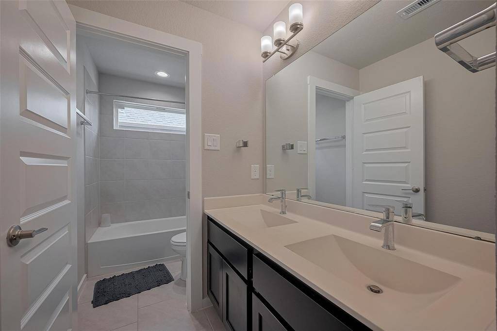 This modern bathroom features a double vanity with sleek faucets, large mirror, and overhead lighting. A separate area includes a shower-tub combo with tiled walls and a window for natural light.