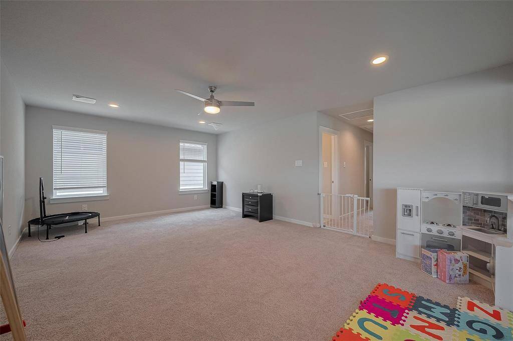 Spacious and bright playroom with neutral carpet, recessed lighting, and a ceiling fan. Features include a children's play area, two windows for natural light, and easy access to adjacent rooms.