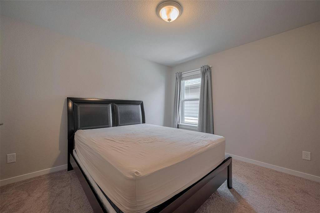 This bedroom features a clean, minimalist design with neutral walls and carpet. It includes a double bed with a modern dark headboard, a window providing natural light, and a central ceiling light fixture.