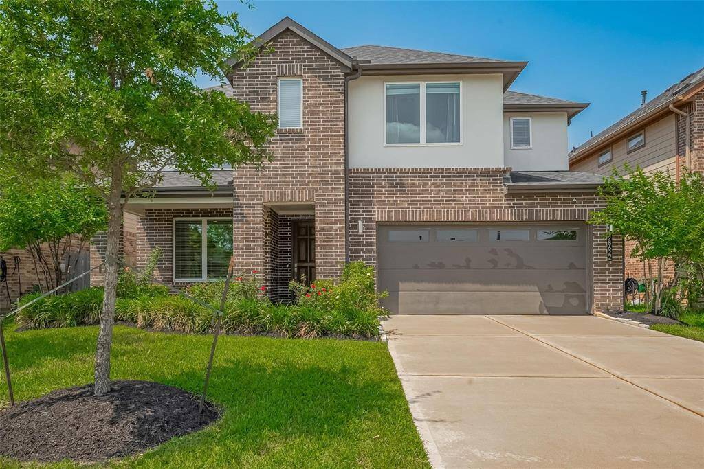 This is a modern two-story brick home with a well-maintained front yard, featuring a tree and landscaped garden beds. It has a three-car tandem garage and a clean driveway, offering great curb appeal.
