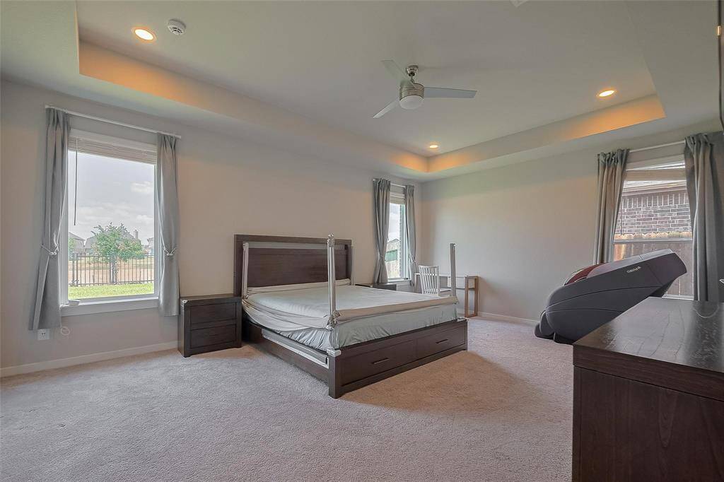 Spacious bedroom with large windows offering natural light, cozy carpet, and recessed ceiling lights. Features a ceiling fan, modern dark wood furniture, and a treadmill. Neutral tones create a serene atmosphere.