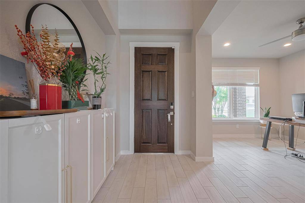 The photo shows a welcoming entryway with a dark wooden door, light wood-style flooring, and neutral walls. A console with decorative items and plants adds charm, while the adjoining room features a large window and ceiling fan, creating a bright and airy atmosphere.