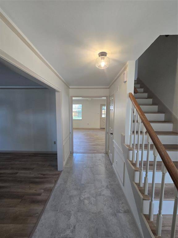 This photo shows a bright entryway featuring tile flooring and a staircase with wooden railings. It leads into a spacious, well-lit room with large windows and wood flooring, providing a welcoming and open feel.
