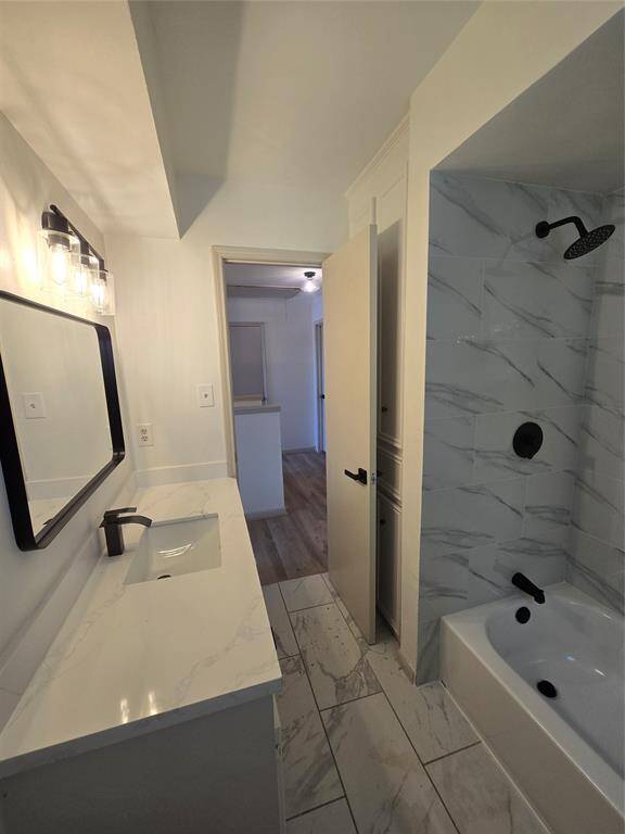 This bathroom features a modern design with a sleek vanity, large mirror, and stylish lighting. The shower area is tiled with a marble pattern, and the fixtures are matte black, complementing the contemporary look. The space opens to a hallway with wood flooring.