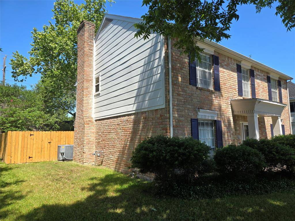 Charming two-story brick home with blue shutters, featuring a side chimney and neatly trimmed bushes. The property has a fenced yard with mature trees, providing shade and privacy.