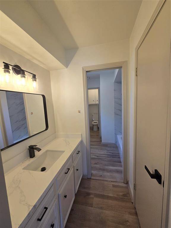 This photo shows a modern bathroom with a long vanity featuring a sleek white countertop and black fixtures. There's a large mirror with stylish lighting above it. The space has wood-style flooring, leading to a separate toilet and shower area.