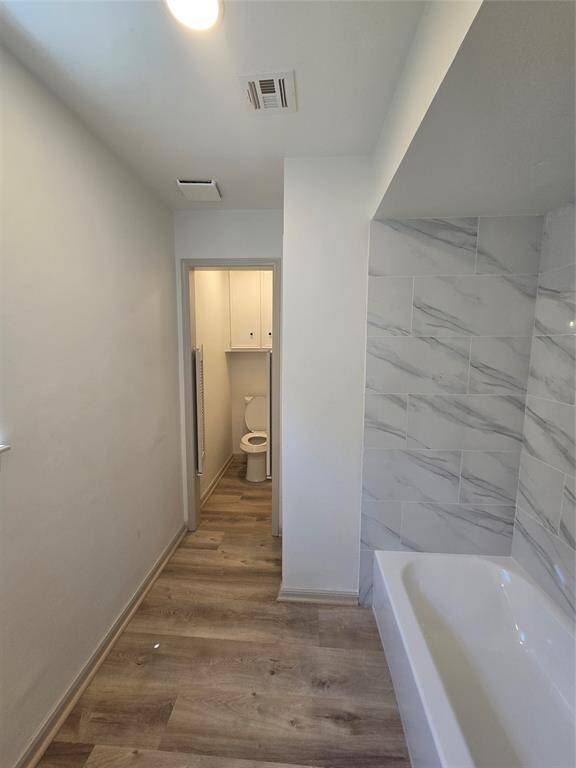 This photo shows a modern bathroom with wood-style flooring, a tiled wall, and a bathtub. There's a separate area for the toilet with cabinetry above for storage. The space is well-lit with recessed lighting.