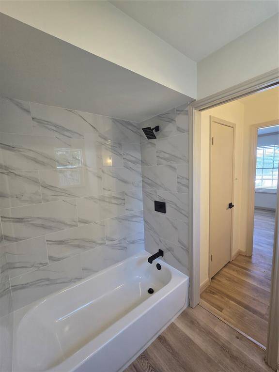 Modern bathroom with a sleek white bathtub, accented by marbled tile walls and matte black fixtures. Adjacent room features light wood flooring and ample natural light from a window.