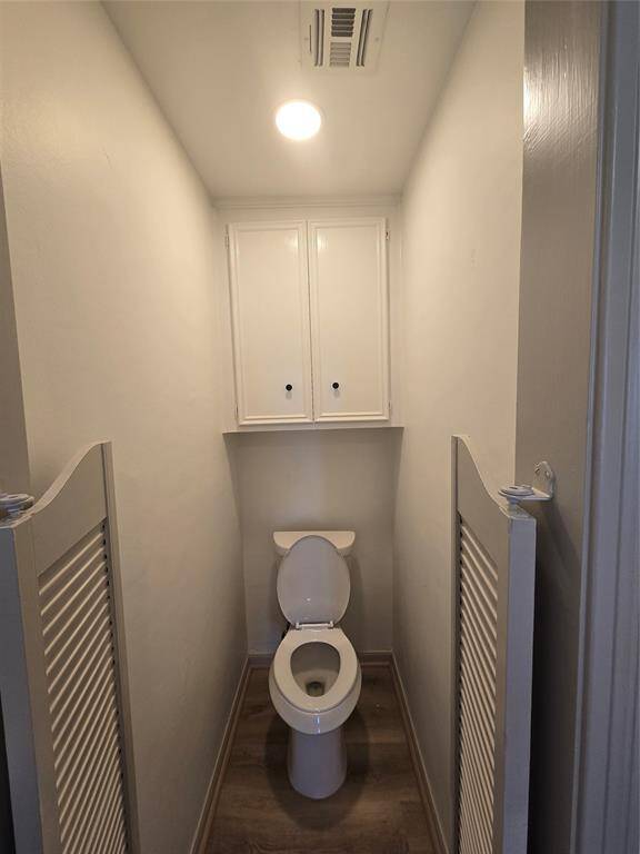This photo shows a small, narrow water closet with a toilet, white walls, and a cabinet above for storage. It features overhead lighting and wood-look flooring.