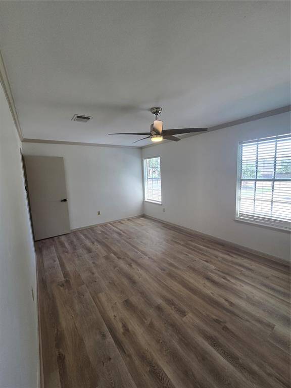 This room features light wood flooring, neutral walls, and ample natural light from two windows. It includes a modern ceiling fan and offers a spacious, airy feel.