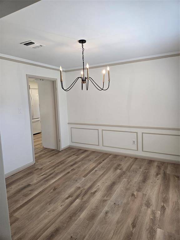 This room features a modern chandelier, light wood flooring, and white walls with decorative molding. It has an open doorway leading to a kitchen area, creating a seamless flow between spaces.