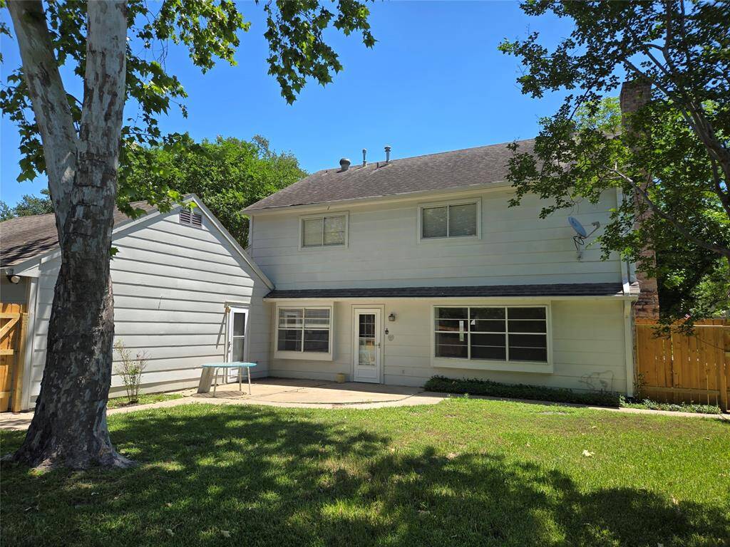 This photo shows the back of a two-story house with light-colored siding, a spacious patio, and a well-maintained lawn. It features mature trees providing shade and a wooden fence for privacy.