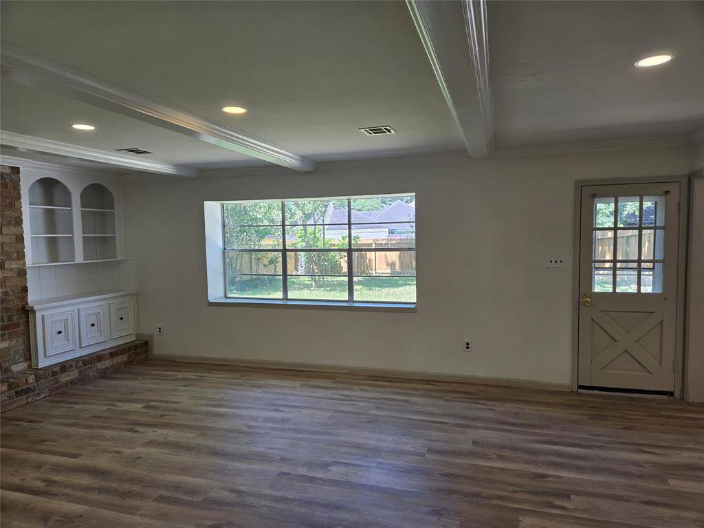 This room features a spacious layout with light hardwood floors, a large window offering natural light, and built-in shelving. There's a brick accent wall and a door leading outside, making it a bright and inviting space.