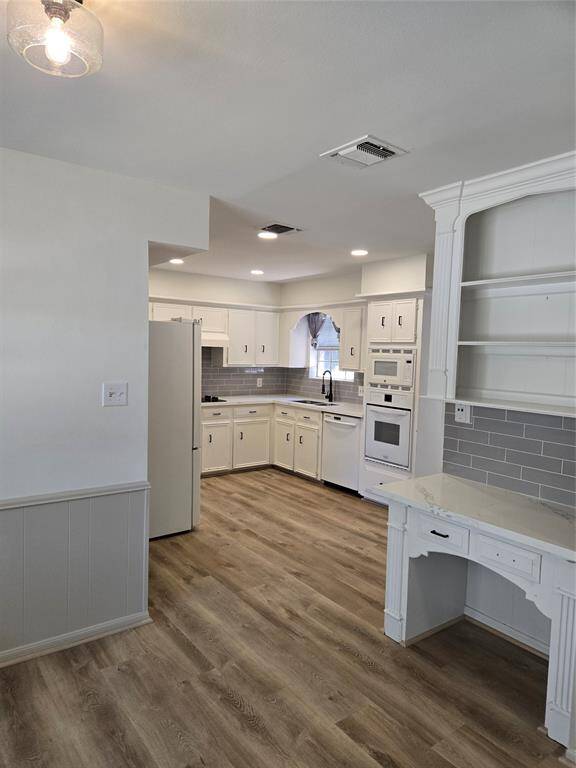 This kitchen features a bright, open layout with modern laminate flooring, white cabinetry, and built-in appliances. A cozy desk area and ample shelving add functionality and charm, while the gray tile backsplash provides a contemporary touch.