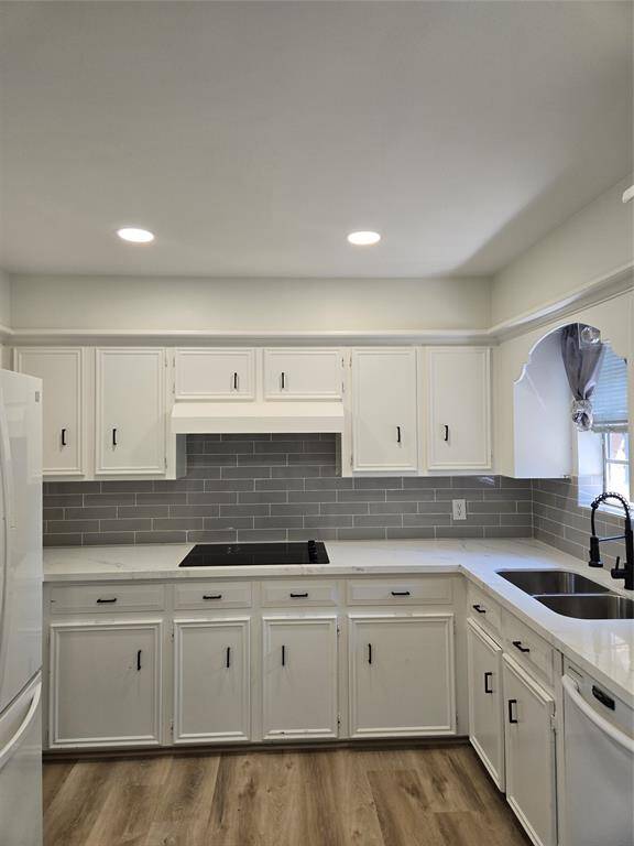 This kitchen features white cabinetry, a sleek gray subway tile backsplash, and modern appliances. It has a built-in stovetop, double sink, and wood-style flooring, offering a clean and contemporary look.