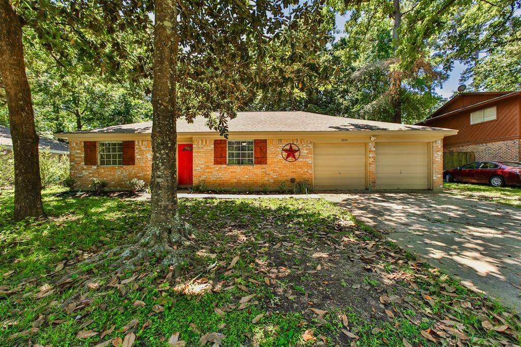Cozy single-story brick home with a charming red front door and two-car garage. Set amidst mature trees, it offers a welcoming, shaded front yard in a serene neighborhood.