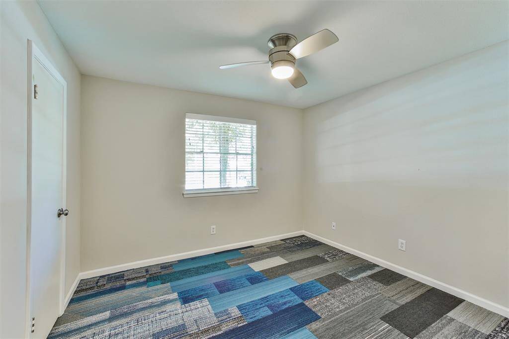 This room features a modern ceiling fan, a window with blinds allowing natural light, neutral wall colors, and a multi-colored patterned carpet, creating a bright and inviting space.