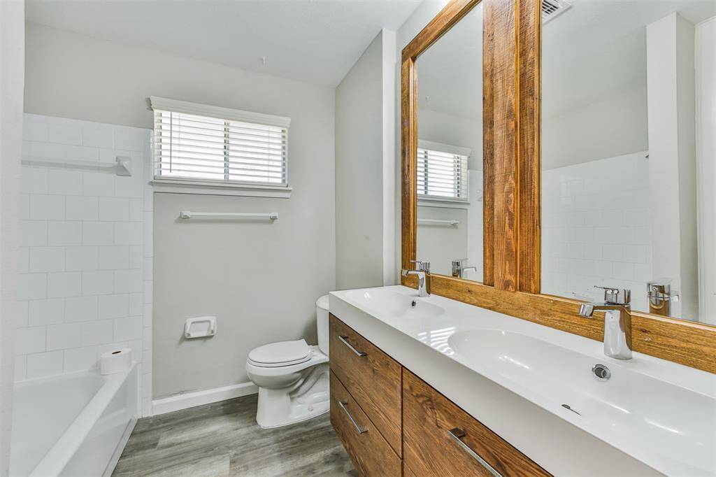 This bathroom features a modern design with a large wooden-framed mirror, dual sinks, and a sleek vanity. It has a bathtub with a tiled surround, a toilet, and natural light from a window with blinds, complemented by light gray walls and wood-look flooring.