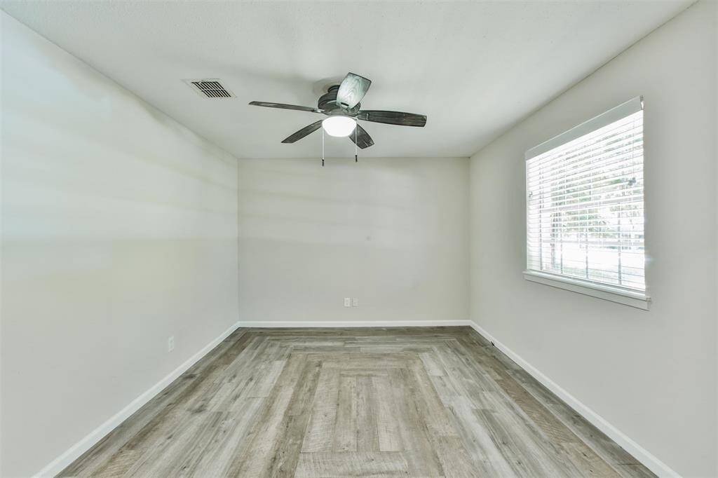 This room features a modern ceiling fan, light gray walls, and wood-style flooring. A large window with blinds provides ample natural light, creating a bright and inviting space.