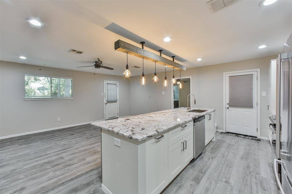 This modern kitchen features a spacious open layout with sleek gray flooring, a large granite island with a built-in sink, and stylish pendant lighting. Ample natural light pours in through a window, enhancing the space's bright, airy feel.