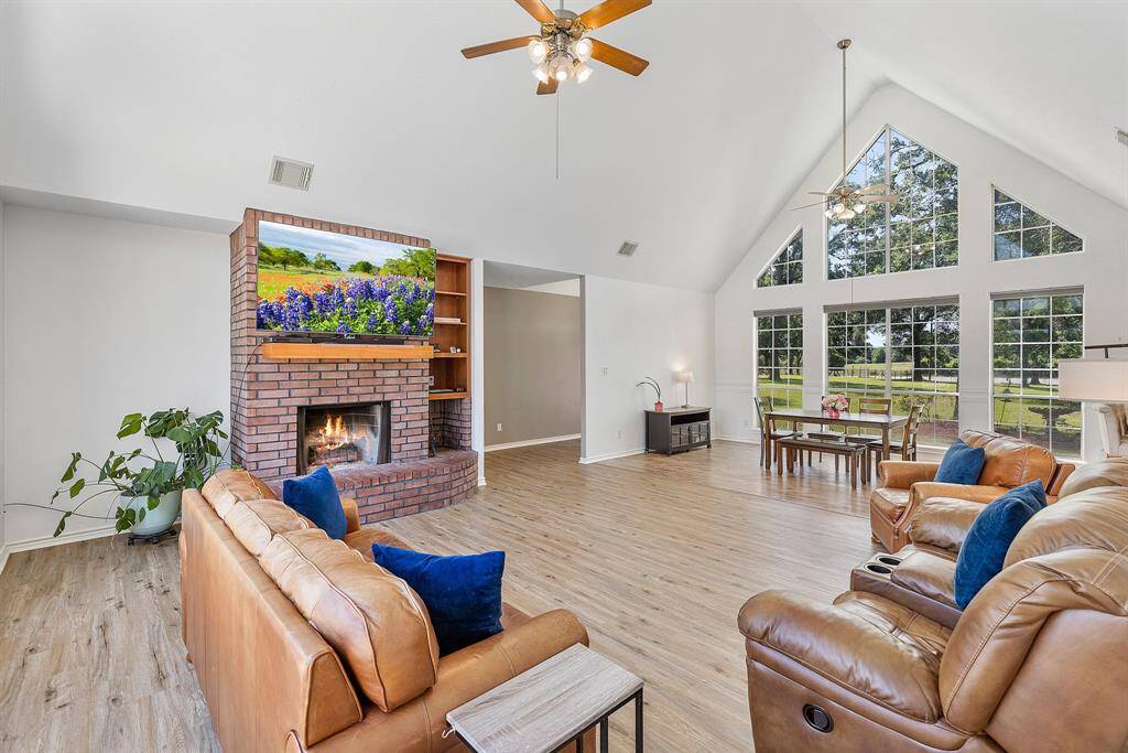 This spacious living room features high vaulted ceilings and large windows that let in plenty of natural light. It includes a cozy brick fireplace, LVT flooring, and comfortable seating with a neutral color palette.