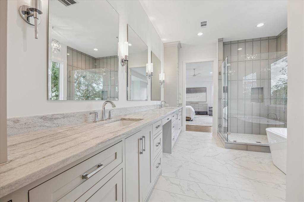 This elegant primary bath features a long double vanity with abundant storage, a frameless glass shower with designer tile, and a freestanding tub positioned to enjoy natural light from large windows.