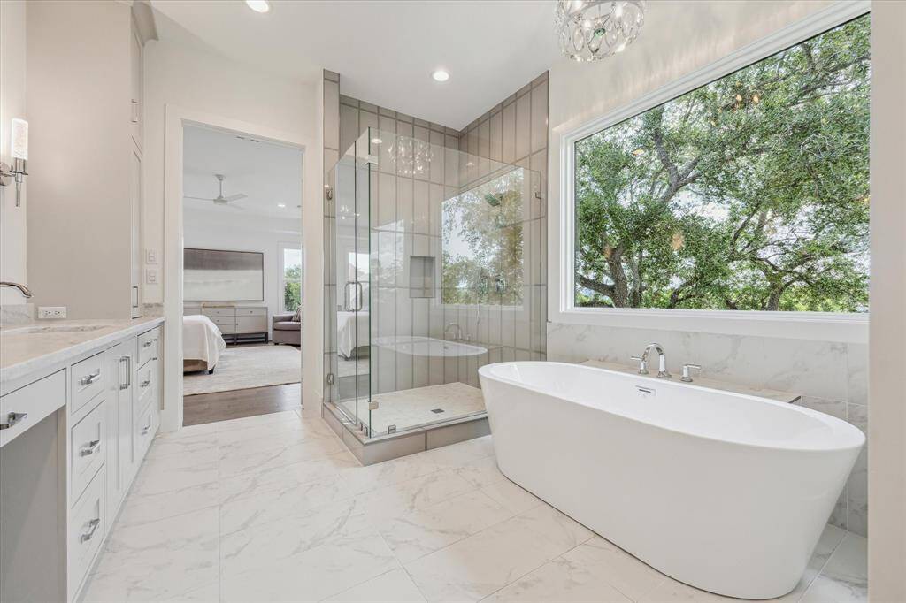 This luxurious primary bath showcases a sleek glass-enclosed shower, a soaking tub set beneath a picture window with lush views, and dual vanities offering generous counter space and storage.