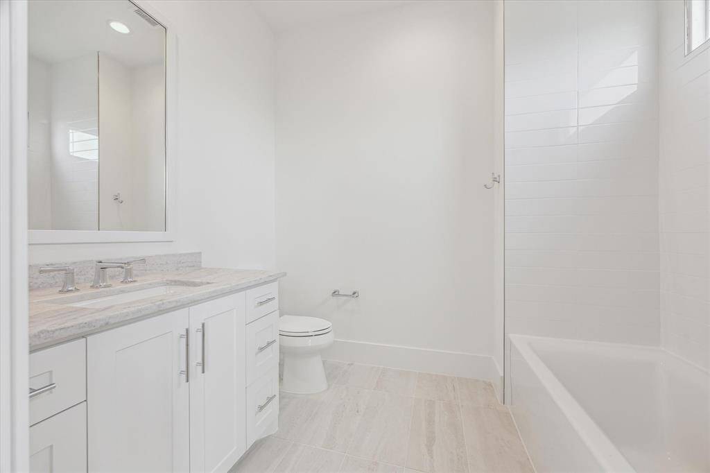 This secondary en suite bath features a sleek vanity with generous counter space, a large mirror, and a combination tub and shower surrounded by clean, modern tilework. A window above the shower brings in soft natural light.