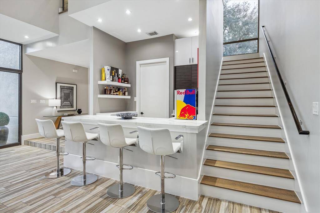 This modern home features an open-concept living area with a stylish bar counter, and built-in shelving. The space is enhanced by sleek flooring, recessed lighting, and a staircase with a large window that brings in natural light.