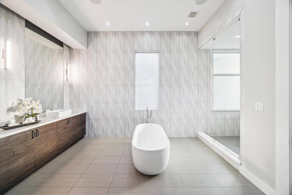 This is a modern bathroom featuring a sleek freestanding tub, textured wall tiles, a double sink vanity with dark wood cabinetry, and a spacious walk-in shower. The minimalist design and neutral tones create a luxurious and serene atmosphere.