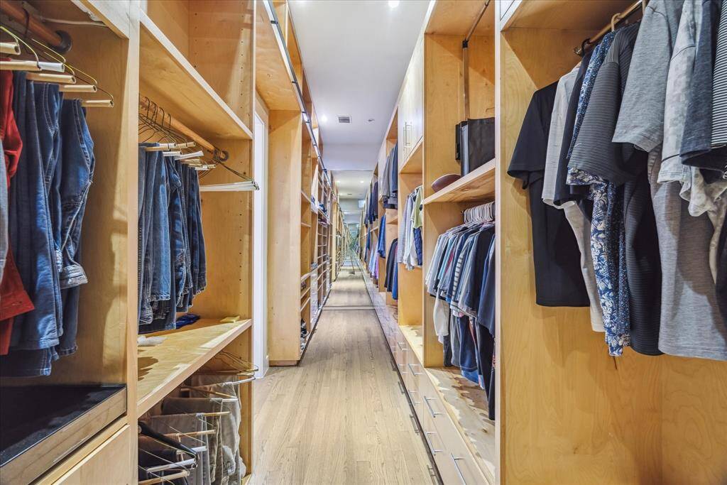 This photo showcases a spacious, well-organized walk-in closet with wooden shelving and ample hanging space for clothes. The closet features a long, narrow layout with a hardwood floor and recessed lighting, providing a bright and functional storage solution.