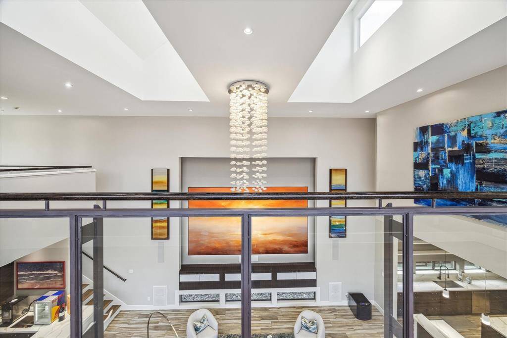 This spacious living area features a modern design with high ceilings, abundant natural light, and a stunning chandelier, while glass railings and open spaces create a sophisticated and airy atmosphere.