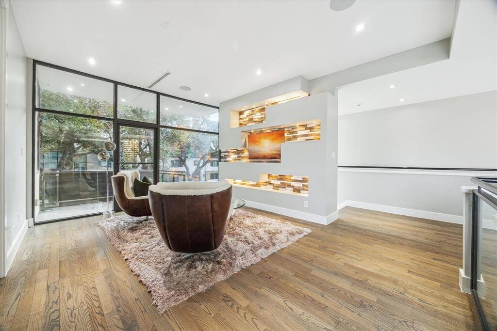 This modern living area features large floor-to-ceiling windows, offering abundant natural light and views of trees. It includes stylish accents like a built-in wall display, and sleek hardwood flooring, creating a warm and inviting space.