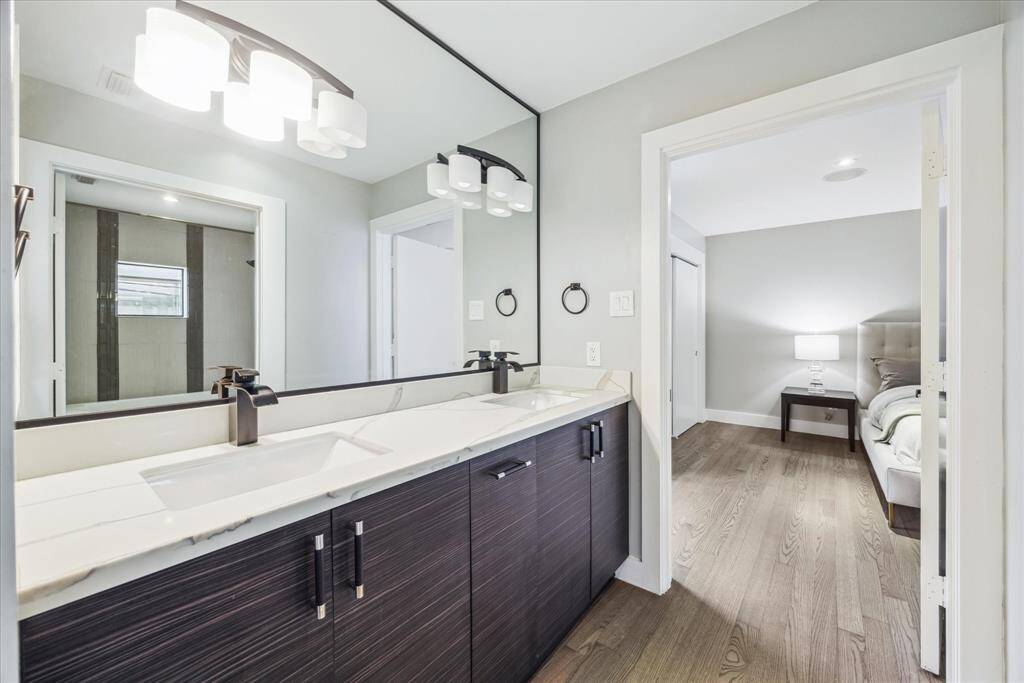 This photo showcases a modern bathroom featuring a double vanity with sleek dark cabinets and large mirrors. The space is well-lit with contemporary fixtures, and an adjoining bedroom is visible with a cozy, inviting setup.